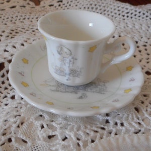 Op de afbeelding: Een wit theekopje en schotelset met een delicaat bloemenontwerp met een meisje met een bloem in haar haar. De schotel heeft een gegolfde rand en is versierd met gele sterren.