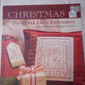 May include: A book titled "Christmas Patchwork Loves Embroidery" with a red, white and green cover. The cover features a red and white striped cushion with the text "Joy to the world" embroidered on it. There is a wrapped gift with a red and white striped ribbon and a string of red and white beads.
