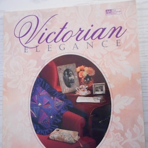 Könnte beinhalten: Buchcover mit dem Titel "Victorian Elegance" in violetter Schrift. Das Cover zeigt ein Foto eines roten Sessels mit einem violetten und blauen Patchwork-Kissen, einer kleinen roten Box und einem Tisch mit einem gerahmten Foto, einer Blumenvase und einer Teetasse und Untertasse.