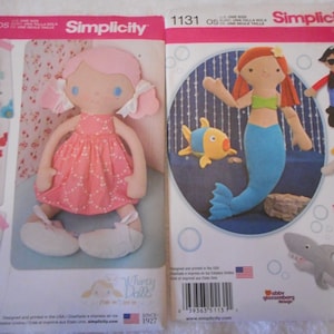 May include: Two Simplicity sewing patterns for making dolls. The first pattern is for a pink and white doll with a pink dress and white shoes. The second pattern is for a mermaid doll with a blue tail and a green top. The patterns are both labeled with the Simplicity logo and the pattern number.