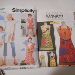 May include: Two sewing pattern packages, one for a dress and one for aprons. The dress pattern is from Simplicity and features a girl wearing a floral print dress. The apron pattern is from McCall's and features three different aprons, one with a shoe design, one with a cat design, and one with a geometric design.