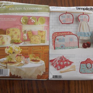 May include: Two sewing pattern books for kitchen accessories. The book on the left is Simplicity pattern number 341 and features a floral design. The book on the right is Simplicity pattern number S8900 and features a red, white, and blue camper design.