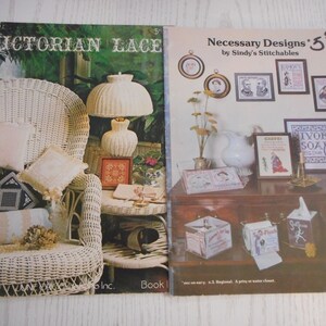 May include: Two cross-stitch pattern books, one titled "Victorian Lace" and the other titled "Necessary Designs" by Sindy's Stitchables. The Victorian Lace book shows a white wicker chair with pillows and a lamp. The Necessary Designs book shows a variety of cross-stitch patterns, including a pitcher, soap, and a jar.