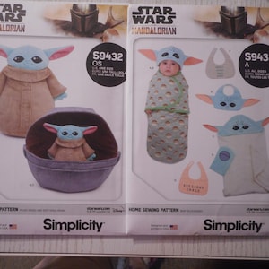 May include: Two Simplicity sewing patterns for making Star Wars Mandalorian baby items. The pattern on the left is for a plush toy and a soft space pram. The pattern on the right is for a baby swaddle, a hooded towel, and a bib. Both patterns feature the character Baby Yoda.