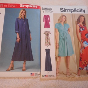 May include: Two Simplicity sewing pattern envelopes. The left envelope is for pattern number S8983 and features a long, blue denim dress. The right envelope is for pattern number S9600 and features three dresses: a pink dress, a floral print dress, and a teal dress.