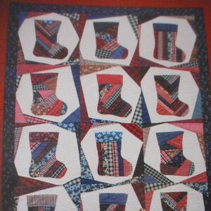 May include: A red, white, and blue patchwork quilt with a Christmas stocking design. The quilt is displayed on a table with two small wooden houses that say "Fruit Shop" and "Quilt Shop".