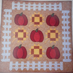 Puede incluir: Una colcha marrón con un borde de valla blanca. La colcha presenta un patrón repetitivo de calabazas rojas y cuadrados amarillos y marrones.