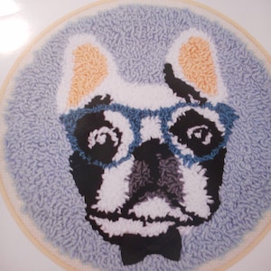 May include: A white and black French bulldog wearing blue glasses and a black bow tie. The dog is on a blue background and is surrounded by a wooden embroidery hoop.