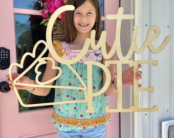 May include: A light-colored wooden cutout sign featuring the words Cutie Pie in a script and block font combination. The design includes a triangular pie slice shape on the left side with two heart-shaped cutouts within the pie slice outline.