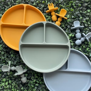 May include: A collection of colorful silicone baby feeding sets. The sets include divided plates in yellow, green, and blue, along with matching forks and spoons. The plates are round with three compartments, perfect for portioning meals.