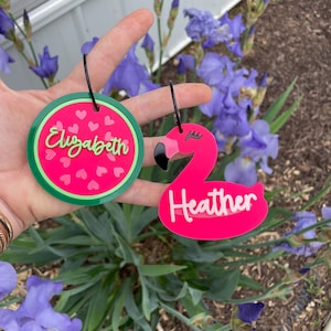 May include: Two personalized acrylic tags. One is watermelon-themed with the name "Elizabeth" in green script. The other is a pink flamingo with the name "Heather" in white script. Both have black loops.