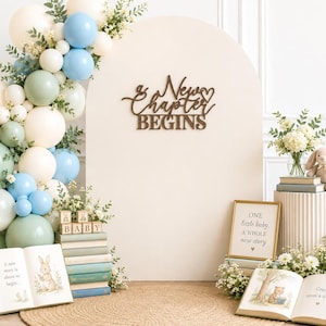 May include: A beige arched backdrop with the wooden text A New Chapter Begins, decorated with a balloon garland in sage green, light blue, and cream. Includes a teddy bear, stacks of books, baby blocks, and open storybooks with illustrations.