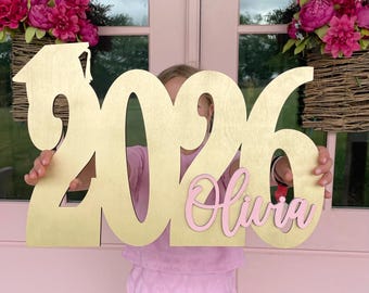 May include: Wooden graduation sign in gold with the year 2026 and the name "Olivia" in pink script. The sign includes a graduation cap. The background features pink doors and hanging baskets of pink flowers.
