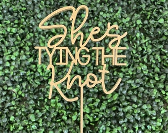 May include: A wooden cake topper with the text "She's Tying the Knot" in a cursive font.