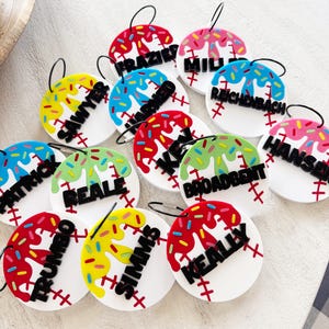 May include: A collection of baseball-themed name tags. Each tag is white with red stitching and a black name. The top of each tag has a colorful, dripped icing design with sprinkles. Each tag has a black loop for hanging.