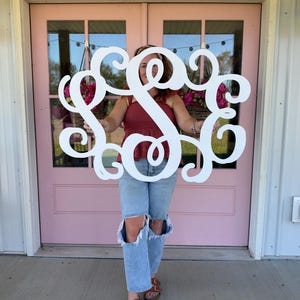 May include: A large, white, custom-cut wooden monogram sign featuring three intertwined script letters with ornate, swirling flourishes. The decorative wall hanging is crafted from a smooth, solid material and displays a classic, elegant cursive font style.
