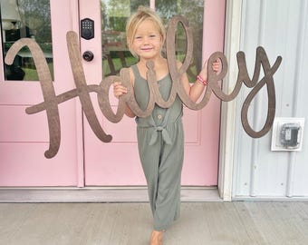 May include: A large, dark brown wooden cutout sign featuring the name Hailey written in a cursive script font. The letters are connected in a single piece, showcasing a smooth, matte finish and a natural wood grain texture throughout the surface.