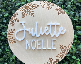 May include: A round wooden sign with the name "Juliette" in white script and "NOELLE" in white capital letters. The sign is decorated with brown snowflake designs around the edge.