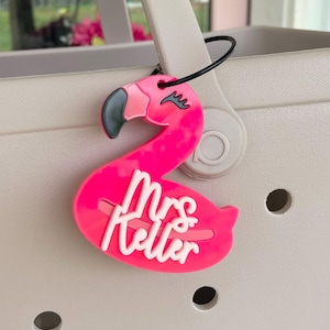 May include: A pink flamingo-shaped bag tag with the name "Mrs. Keller" in white script. The flamingo has a black eye and a black loop for attaching to a bag. The tag is hanging from a light beige tote bag.