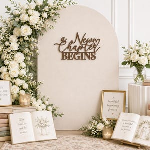 May include: A beige arched backdrop featuring a dark brown wooden sign with the text A New Chapter Begins. The display includes stacks of books, white floral arrangements, candles, and framed signs with text including The best is yet to come.
