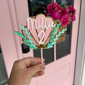May include: A pink and gold seashell-shaped cake topper with the name "Millie" and the number "2" in white. The topper has green accents and is held by a pink stick. The background includes a pink door and a hanging basket of pink flowers.