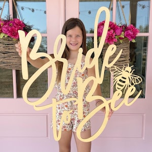May include: A wooden sign that says "Bride to Bee" in a cursive font, with a bee silhouette. The sign is held by a person wearing a floral romper. Pink door and floral baskets in the background.