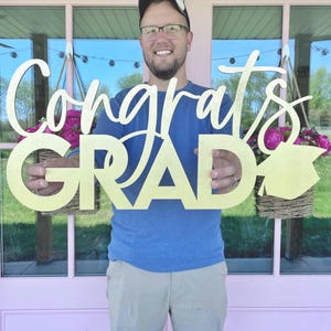 May include: A gold-colored wooden sign that says "Congrats GRAD" in a script font. The sign is held by a person in front of a pink door with hanging baskets of pink flowers. The person is wearing a blue shirt, glasses, and a black hat.