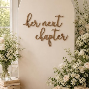 May include: A tall, arched, cream-colored backdrop featuring the brown, cursive, three-dimensional wooden text "her next chapter" mounted in the center. The display is surrounded by white floral arrangements and stacks of vintage books.