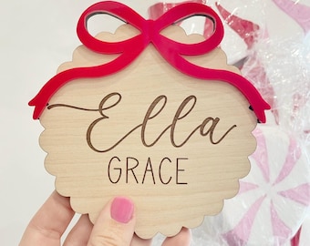 May include: A wooden ornament with the name "Ella Grace" engraved in a script font. A bright red bow is attached to the top of the ornament. The ornament has a scalloped edge. In the background are white and pink peppermint candies.