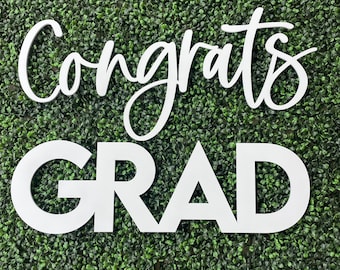 Pode incluir: Uma placa branca com a palavra Congrats em uma fonte cursiva, posicionada acima da palavra GRAD em uma fonte sem serifa em negrito e maiúsculas. As letras são cortadas individualmente e apresentam um acabamento branco.