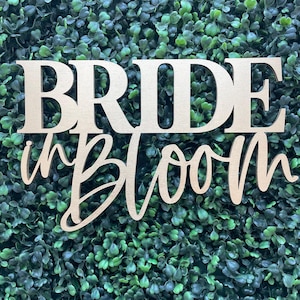 May include: A light-colored wooden sign with the text "BRIDE in Bloom" in a combination of bold block lettering and a cursive script font, positioned against a dense background of small, vibrant green artificial leaves and foliage.