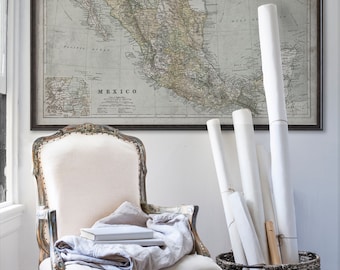 May include: A vintage map of Mexico in a black frame hangs on a white wall above a white chair with a linen throw and books. A wicker basket holds rolled up white paper.
