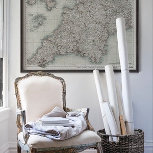 May include: A vintage map of the South West of England in a black frame hangs on a white wall above a white upholstered chair with a linen throw and books.  A wicker basket holds rolled up white paper.