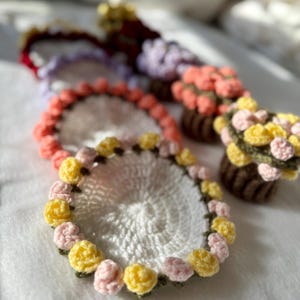 May include: Crocheted coasters and small flower pot decorations. The coasters are white with a floral border of yellow and pink flowers. The flower pots are brown with pink and yellow flowers. The items are on a white, textured surface.