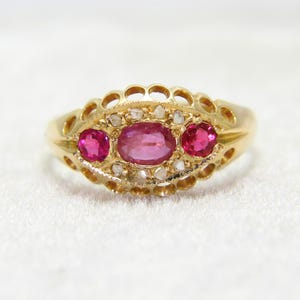 May include: A gold ring featuring a central oval-cut pink gemstone flanked by two smaller round-cut pink gemstones. The setting is surrounded by small, rough-cut white stones and a decorative, openwork scalloped gold border along the band.