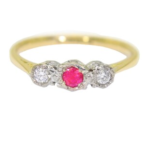May include: A gold ring featuring a central round-cut vibrant pink gemstone flanked by two smaller round-cut clear white diamonds. The stones are set in a silver-toned metal setting that contrasts with the smooth, polished yellow gold band.