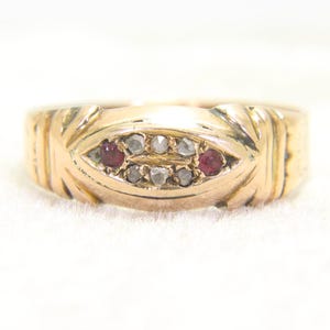 May include: A gold ring featuring an elongated, marquise-shaped center setting. The setting contains two small, round, deep red stones positioned at the far ends, flanked by six small, rough-cut, translucent grey stones arranged in two rows.