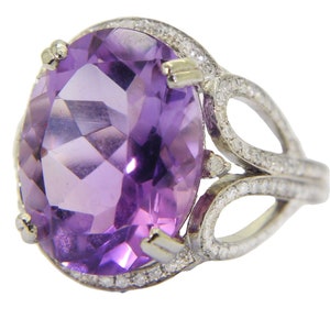 Könnte beinhalten: Ein silberfarbener Ring mit einem großen, oval geschliffenen violetten Amethyst, der von vier Krappen gehalten wird. Die Ringschiene und die Fassung um den zentralen Stein sind mit zahlreichen kleinen, klaren, rund geschliffenen weißen Steinen besetzt.