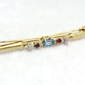 Puede incluir: Un broche de barra de oro con una topacio azul, rubíes rojos y diamantes blancos. El broche está engastado en una barra de oro con un extremo puntiagudo.