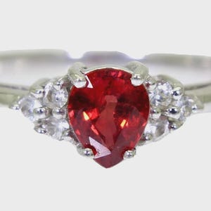 Peut inclure: Une bague en métal blanc avec une pierre rouge en forme de poire et des petites pierres blanches de chaque côté.