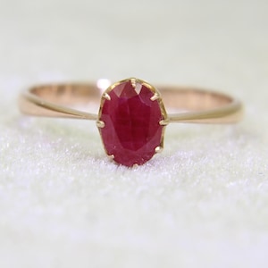 Puede incluir: Un anillo de oro con una piedra preciosa de rubí rojo intenso de corte ovalado en el centro, sujeta por seis pequeñas garras de metal. La banda del anillo es lisa y se estrecha ligeramente donde se une al engaste de la piedra roja facetada.