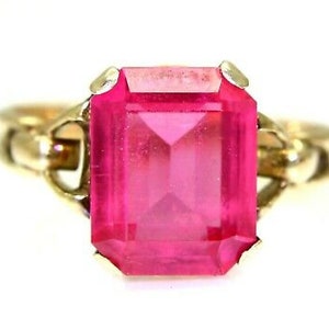 Könnte beinhalten: Ein goldener Ring mit einem großen, leuchtend pinkfarbenen, smaragdgeschliffenen Edelstein.