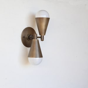 May include: A wall-mounted light fixture with a bronze finish. The fixture has a circular base, a connecting arm, and two cone-shaped shades. Each shade holds a white, globe-shaped light bulb. The fixture is mounted on a white wall.