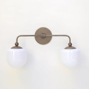 May include: A brass wall sconce with two white frosted glass shades. The sconce has a curved arm and a round back plate.