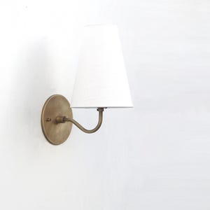 May include: A wall-mounted lamp with a white lampshade and a brass-colored metal base. The lamp has a curved arm and a round backplate. The lampshade is a classic tapered shape.
