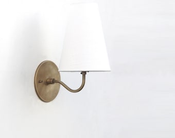 May include: A wall-mounted lamp with a white lampshade and a brass-colored metal base. The lamp has a curved arm and a round backplate. The lampshade is a classic tapered shape.