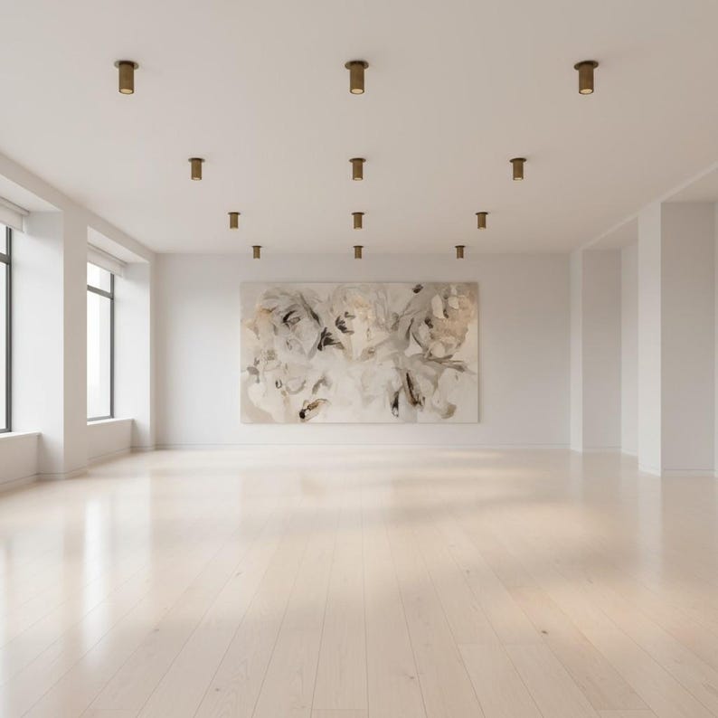 May include: A spacious, empty room with light-colored wooden flooring and white walls. A large, rectangular abstract painting with beige, white, and grey tones hangs on the back wall. The ceiling features multiple cylindrical bronze spotlights.
