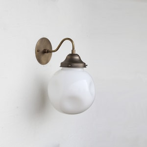Puede incluir: Una lámpara de pared de latón con una pantalla de vidrio blanco texturizado en forma de globo. La lámpara tiene un brazo curvo y una placa trasera redonda.