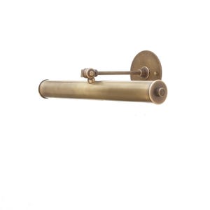 May include: A brass wall-mounted picture light with a cylindrical shade and a round wall plate. The light is adjustable and can be tilted up or down.