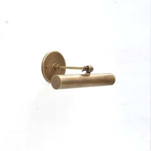 Puede incluir: Una lámpara de pared para cuadros con cabezal cilíndrico de latón y placa trasera circular. La lámpara cuenta con un brazo horizontal delgado conectado al tubo de luz mediante un pequeño mecanismo de bisagra ajustable, acabado en latón.